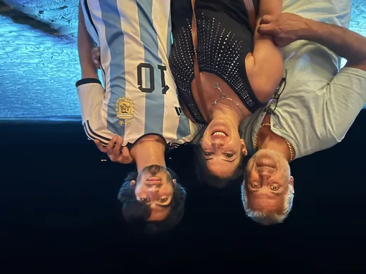 Three people smiling together at night on a beach, with the young man wearing an Argentina soccer jersey